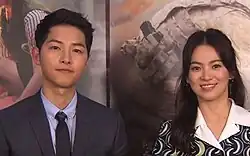 Actors Song Joong-ki and Song Hye-kyo pose side-by-side in front of a promotional backdrop for "Descendants of the Sun"