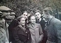 Black and white photo of a Soviet soldier talking to civilians in a village setting.
