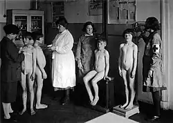 four nude children receiving a medical exam at school from four women
