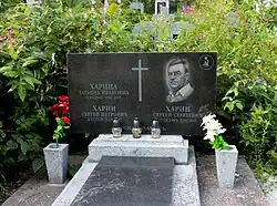 A dark stone gravestone with an engraved cross and portrait stands in a leafy cemetery, with candles and flowers placed in front. Overgrown greenery and flowers surround it, creating a quiet, solemn scene.