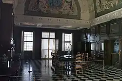 Interior of the Monplaisir Palace showing a formal hall with a long table and chairs, checkered tile floor, tall windows, and an ornate painted ceiling.