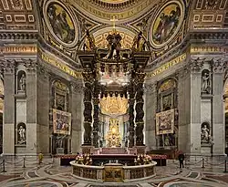 Photo shows the baldachin standing in the centre of the church, viewed looking towards the nave. There is an altar beneath it which has a red and gold frontal cloth decorated with large crosses.