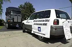 An SUV (RAV4 EV) with a poster that reads "GM Don't Crush the EV1s! Zero Emissions NOW!