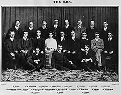Group photograph of a seated woman in Victorian dress surrounded by 19 suited men.