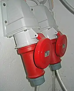 In the foreground, a downward-angled industrial wall socket with a red cap, mounted to a wall; a red plug is inserted into it. In the background, another similar connection.