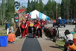 Traditional Tlingit dancers performing outdoors at the Teslin Tlingit Heritage Centre, beside a totem pole.
