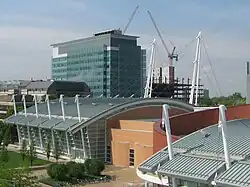 View from above of a building with white poles sticking up from its sides and the cables connecting them. A taller building is in the background and a connected brick building is visible on its right.