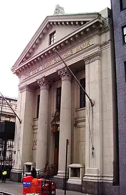 The main facade of the building on Eighth Avenue. The facade is designed as a classical portico and is divided vertically into three bays, with two fluted columns on the inside and a pair of paneled pilasters with antae on the outside. These support an entablature and a triangular pediment with a window. Metal letters with the words "The New York Savings Bank" are placed on the entablature.