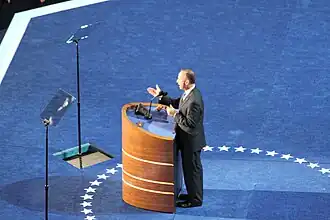 (A) The first three teleprompters: A monitor screen partly embedded in the lectern's desktop displays the scrolling text of the speech in synchrony with two screens embedded in the podium floor. They are on either side of the speaker, reflected by the angled glass teleprompters above them.