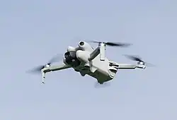 A grey quadcopter seen from a low angle against a pale blue sky, featuring a front-mounted camera and four spinning propellers.