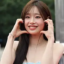 A young Asian woman with long brown hair and a fringe wearing a blue dress grinning widely with her fingers and thumbs on each of her cheeks as if forming a heart.