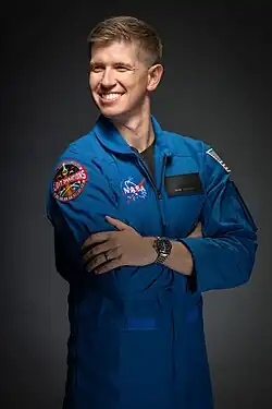 NASA astronaut Adam Fuhrmann, a man in a blue flight suit, is in front of a dark background. He smiles and looks to the left.