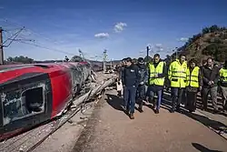 A photo of a red high speed train on its side while men in yellow safety vests inspect it.