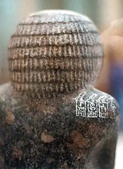 Close-up of the back of the head and shoulder of a granite statue showing white inscribed hieroglyphs on a reddish-brown background