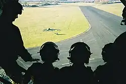 Five troops wearing helmets peer out the open door of an aircraft. Airport taxiways and green lawns appear below them.