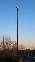 A video camera mounted on a tall pole on the side of a roadway. The camera is not pointing at the roadway visible at the bottom-right of the picture, but to the left.