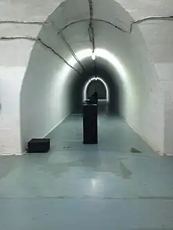 A long, dimly lit concrete tunnel with pipes running along the ceiling and a rail track on the floor.
