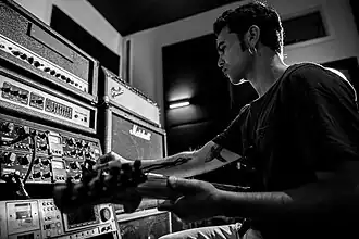 black-and-white image of a seated Arli Liberman from profile, holding an electric guitar and fiddling with a control board with his right hand