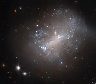 A galaxy fills up most of the frame from the right. It is fuzzy and diffuse, but made up of numerous tiny stars. In the core, the stars merge into a glowing bar shape. The gas and stars in the galaxy vary between warm and cool colours. They are spread over a large area, the colours mixing like clouds. The glow of the galaxy fades into a black background, with a few stars and small, distant galaxies.