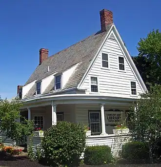 View of side of house, with flowers and shrubbery in front and a sign saying "Schenectady County Public Library, Scotia Branch" on wall behind porch.