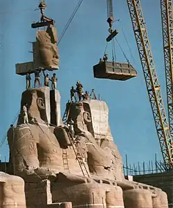 A huge rock sculpture is being dismantled by cranes. The sculpture consists of two large, seated humans.