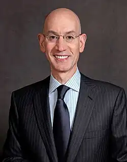 A thin, bald man in a dark pinstripe suit, wearing glasses, striped shirt and dark blue tie against a gray background