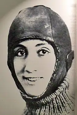 A sepia-tinged black-and-white photograph showing the head of a smiling woman. All but her face is under a leathery covering