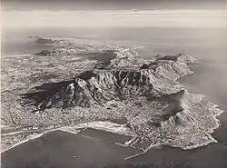 Black-and-white photograph of a city, taken from the air. The city is situated between an ocean and a large flat mountain range.