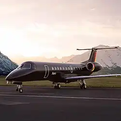 A matte black Aero ERJ-135 jet on the runway at sunset, backed by snow-capped mountains.