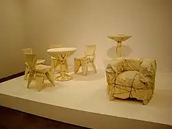 Four cafe chairs and a table, a high table, and an upholstered chair are wrapped in fabric and rope.