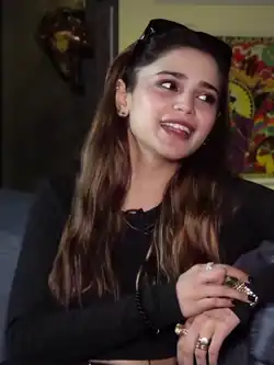A woman, with rings, bracelet, earrings, and a lavalier microphone, having tattoos on her hands and a sunglasses over her head, smiling to her left, showing her right hand's bent middle finger, and talking to the right side of the camera.