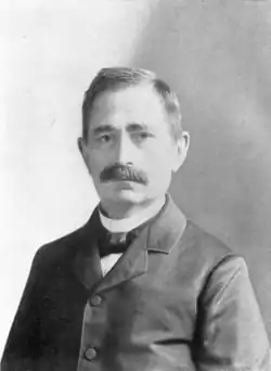 An elderly mustachioed man in a suit with a bowtie