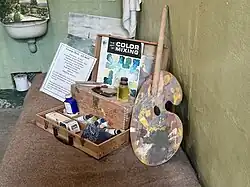 An artist’s paint box with brushes, paints, and a palette sits on a cot, alongside a sign about George Heck, an Alcatraz inmate who painted during his time there.