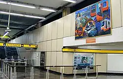 Interior of the station concourse with wall panels, metal railings, directional signage, and a large colorful mural mounted high on the wall above the passageway.