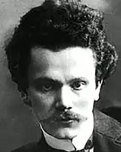 A cropped version of a dramatic black and white studio portrait of a clean-shaven young man in pre-1914 three-piece suit and wing collar, with dark hair, bouffant on top. In the original picture he is seated, leaning on a desk, with a fixed gaze to camera, with striking, dark eyes.