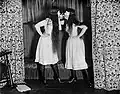 A black and white photograph of two women, both wearing white knee-length dresses, black tights, black shoes, and white Venetian masks covering their eyes and nose. The women have cigarettes in their mouths and are holding the tips to each other.