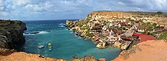 A cluster of brightly painted wooden houses sits beside a turquoise bay, nestled beneath rugged cliffs with dramatic views of the open sea