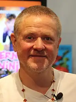 close-up of Anders Matthesen wearing a white t-shirt with a lapel microphone, staring directly at camera with a smile