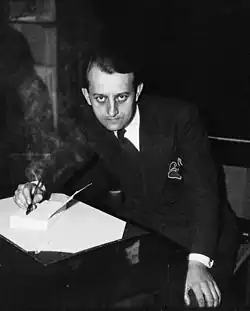 A seated, young, dark-haired man in a suit, photographed from above, is shown writing on a sheet of paper on a desk while staring straight up at the camera.