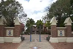A brick wall with a gap in the middle. Through the gap one can see a statue at the bottom of a palm-lined walkway.