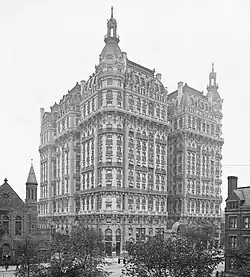 A black-and-white photograph shows a grand, ornate multi-story Beaux-Arts style building with elaborate facades, towers, and decorative details.