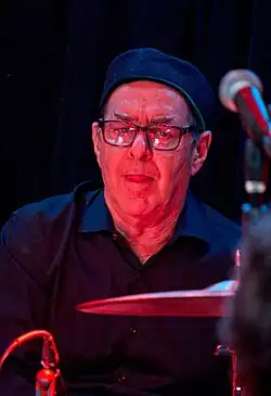 Portrait of a man (Anton Fig) wearing dark glasses and a purple shirt, playing a cymbal that appears in the foreground.