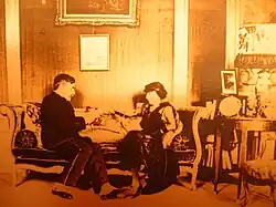 A man and a woman seated on a sofa in conversation in a richly furnished salon. The man, wearing gaiters, reads a manuscript.