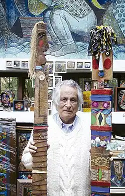 An elderly man in a white sweater stands between two tall sculptures made of painted wood, fabric, and found objects in a colorful, art-filled studio