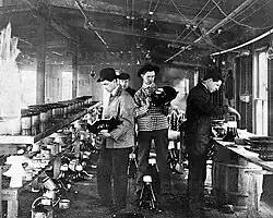 Four men working in a rustically equipped laboratory. Benches covered with equipment are on both sides. One bench is supported by a wooden barrel. Wires can be seen suspended from the ceiling.