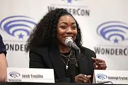 Hairston is sitting at a table speaking into a microphone. She is a Black woman with brown skin and long curly black hair.