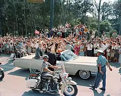 In an open-topped convertible with a motorcycle escort. Very large crowd, many taking photographs.