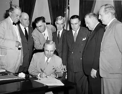 A man in a suit is seated at a desk, signing a document. Seven men in suits gather around him.