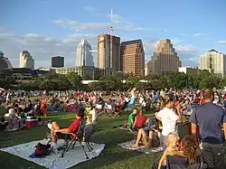 Slightly elevated image taken during the evening of a crowd of people relaxing in a park, with a skyline visible in the background.