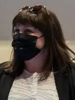 A photograph of a brown-haired woman with sunglasses resting on top of her head and a black face mask covering her nose and mouth